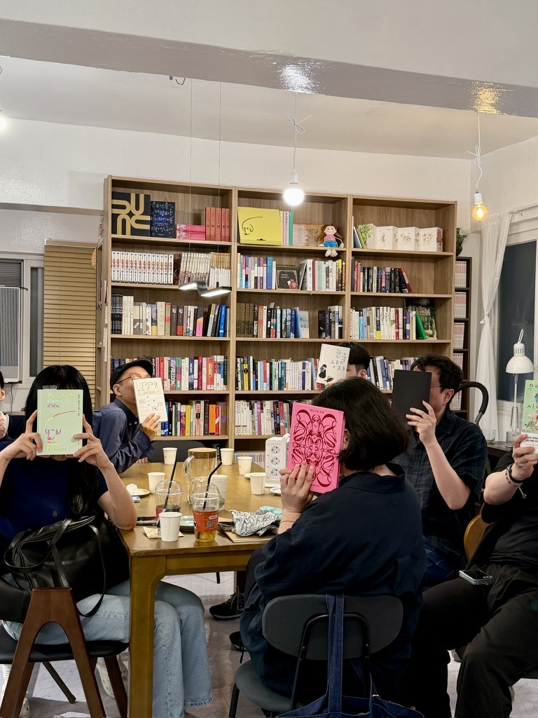 서가 앞 긴 테이블에 둘러앉아 저마다 읽은 책으로 얼굴을 가린 모임 참가자들